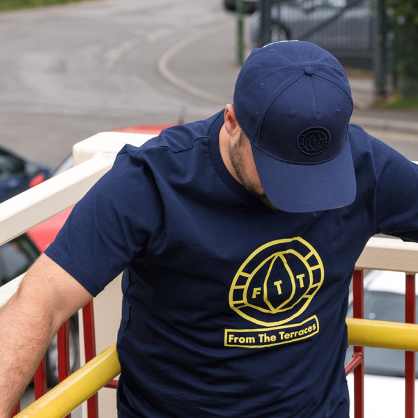 Man wearing a navy blue t-shirt with a yellow logo and text, leaning against a railing.