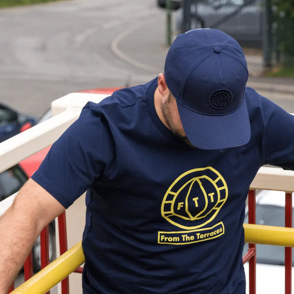 Man wearing a navy blue t-shirt with a yellow logo and text, leaning against a railing.