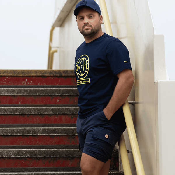 Man wearing a navy t-shirt with a yellow logo and shorts, leaning against a staircase.