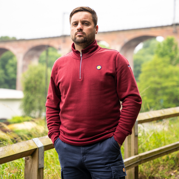 Man wearing a red fleece standing outdoors with a bridge and greenery in the background