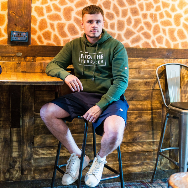 Man sitting on a stool in a rustic wooden bar setting wearing a green hoodie with text.
