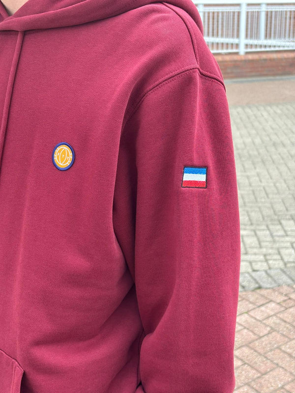 Maroon hoodie with a logo on the chest and another on the sleeve, outdoors.