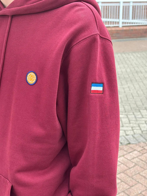 Maroon hoodie with a logo on the chest and another on the sleeve, outdoors.