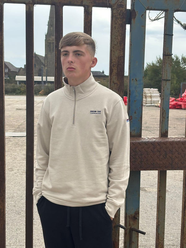 Person wearing a beige pullover with a logo, standing in front of a metal gate.
