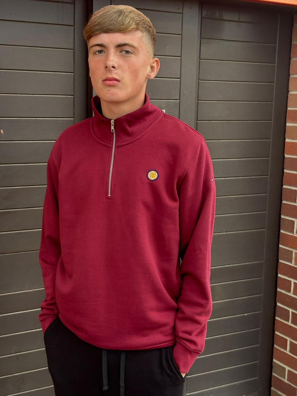 Person wearing a red pullover with a logo, standing against a brick wall.