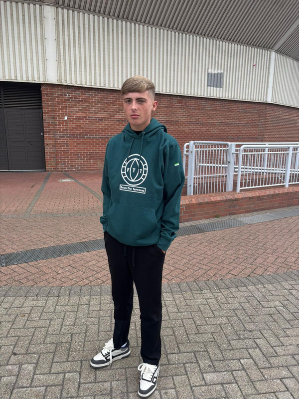 Person wearing a green hoodie with a logo standing on a brick pavement.