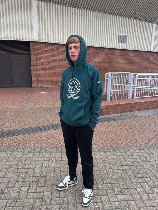 Person wearing a green hoodie with a logo, standing on a paved area with a building in the background.
