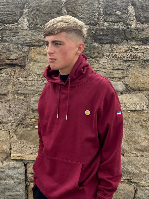 Person wearing a maroon hoodie with logos against a stone wall