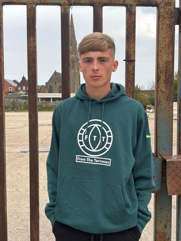 Person wearing a teal hoodie with a logo in front of a rusty metal gate.