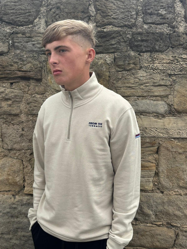 Person wearing a beige pullover with a logo against a stone wall