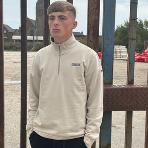Person wearing a beige pullover with a logo, standing in front of a metal gate.