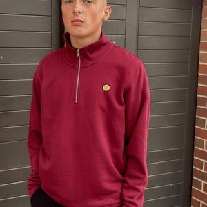 Person wearing a red pullover with a logo, standing against a brick wall.