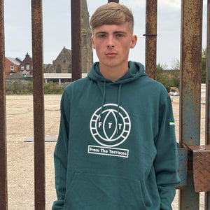 Person wearing a teal hoodie with a logo in front of a rusty metal gate.